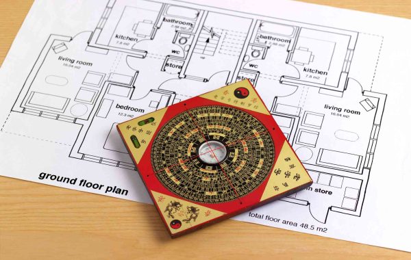 A Chinese feng shui compass with intricate markings rests on an architectural floor plan. The layout includes labeled rooms. The scene conveys balance.